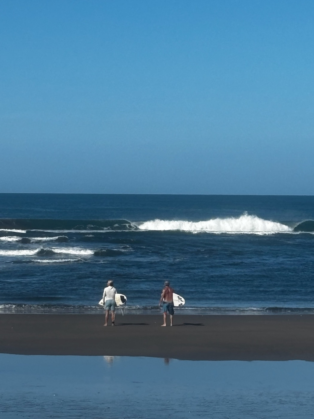 Two surfers walking across dark sand toward the water, boards under their arms, under a clear blue sky.